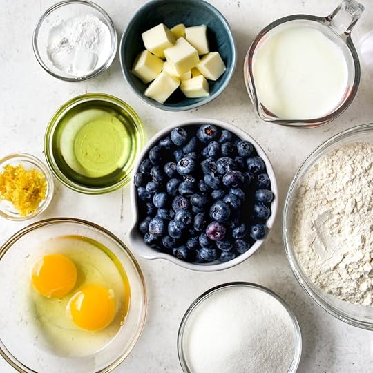An overhead shot of various ingredients for a lemon blueberry loaf cake, arranged in clear glass bowls and small blue bowls on a white countertop. Ingredients include: a bowl of fresh blueberries, two eggs, lemon zest, oil, flour, sugar, baking powder, cubed butter, and a measuring cup of milk.