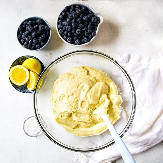 An overhead, close-up shot of a clear glass bowl containing a thick, pale yellow cake batter, with a large pile of fresh, dark blue blueberries poured onto one side of the batter. A white spatula is positioned to begin folding the blueberries into the mix.
