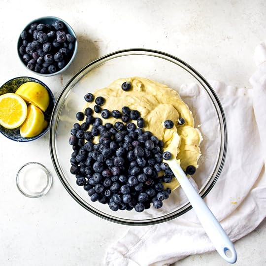 An overhead, close-up shot of a clear glass bowl containing a thick, pale yellow cake batter, with a large pile of fresh, dark blue blueberries poured onto one side of the batter. A white spatula is positioned to begin folding the blueberries into the mix.