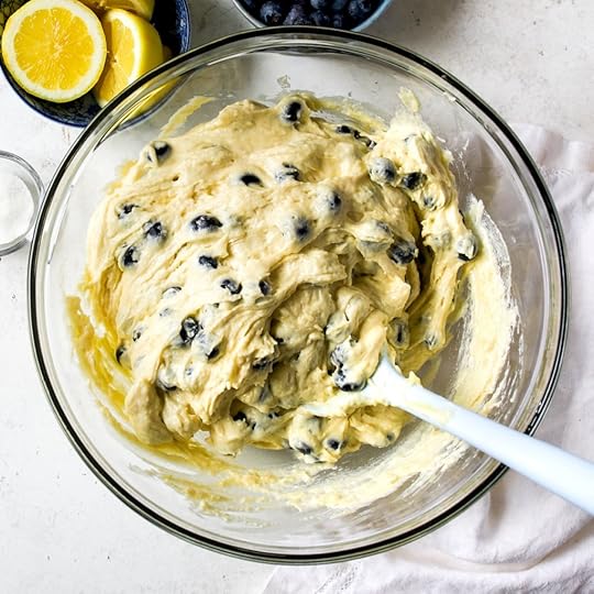 An overhead, close-up shot of a clear glass bowl filled with thick, pale yellow cake batter, generously mixed with dark blue blueberries. A white spatula is visible, having just folded the berries into the batter, which is spattered on the inside of the bowl.