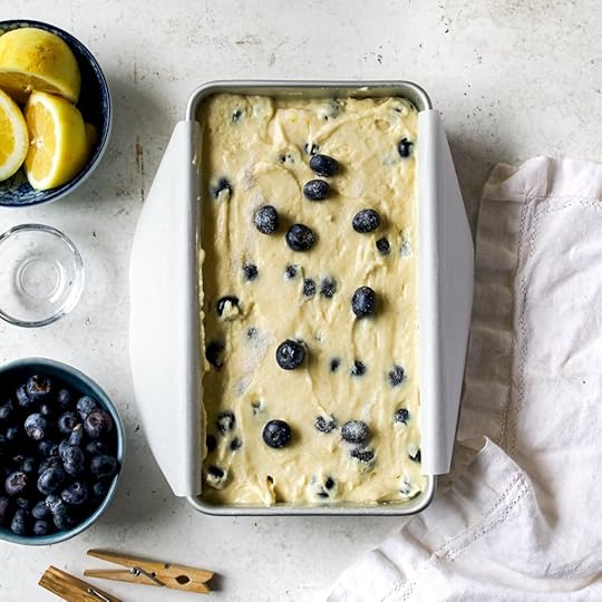 An overhead shot of a stainless steel loaf pan, lined with white parchment paper, filled with a pale yellow lemon blueberry cake batter. Dark blue blueberries are visible throughout the smooth batter, which is ready for baking.