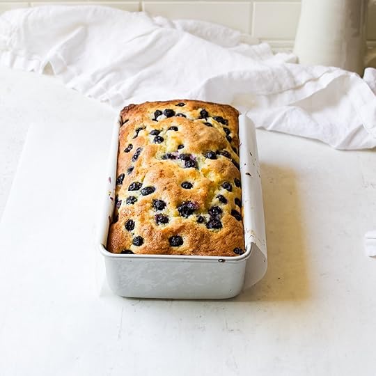 A newly baked, golden-brown blueberry loaf in a silver loaf pan, lined with white parchment paper, sitting on a white counter. The top of the loaf is risen and dotted with dark blueberries. In the soft-focus background, a white ruffled linen napkin and a white pitcher are visible against a white tiled wall.