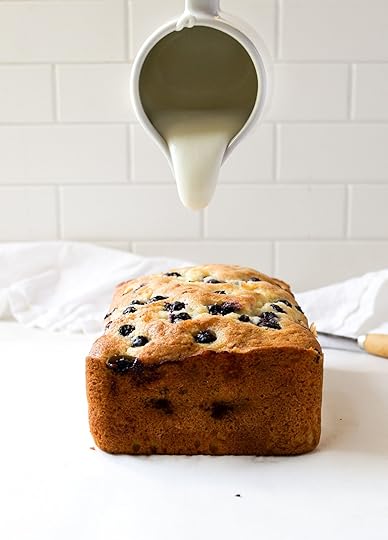 A freshly baked, golden-brown blueberry loaf sitting upright on a white surface, with a white pitcher pouring a stream of white glaze onto its visible, blueberry-studded top. In the blurred background, a white tiled wall is visible, along with a white cloth and a knife handle.