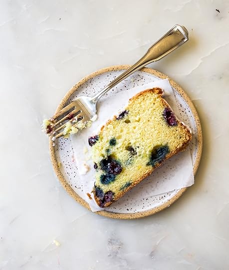 An overhead, close-up shot of a partially eaten slice of blueberry lemon loaf cake, showing its golden-yellow crumb studded with dark blueberries and remnants of white glaze, on a speckled ceramic plate with a textured rim. A silver fork with a bite of cake on its tines rests next to the slice, all on a light marble surface.