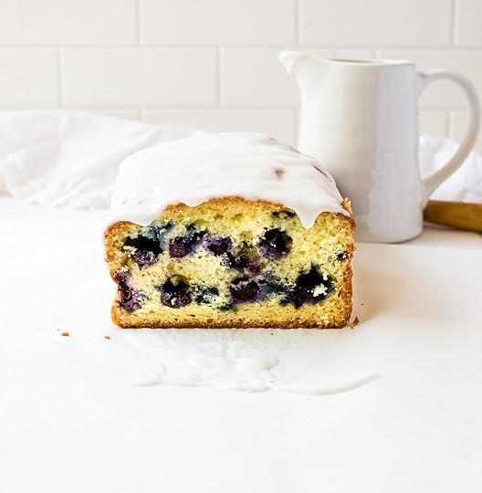 A straight-on, eye-level shot of a glazed blueberry lemon loaf, showing its golden-brown crust and moist interior studded with purple blueberries. The loaf is sitting on a white surface with drips of white glaze, and in the soft-focus background, a white pitcher and a white linen cloth are visible against a white subway tile wall.