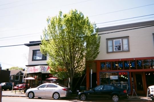 Photo, taken on a bright spring day with an old disposable camera, of a friendly neighbourhood street: cars parked in the shade of a tree growing on the sidewalk in front of Phinney Books and its next-door neighbour, the 74th Street Alehouse.
