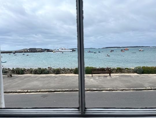 View through a wood-framed window over a bay with fishing and other small boats moored. In the background the large Scillonian ferry is moored at teh end of the stone quay.