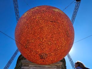 Luke Jerram, Artist’s, Helios at Ickworth. Inflatable orange ball set against a classical portico with hints of the gantry holding it aloft and behind it an azure sky.