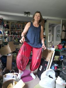 Picture of messy room containing a middle age woman wearing a floaty dark blue vest top with red pantaloons. She is holding the pantaloons out to show how voluminous they are and grinning inanely.