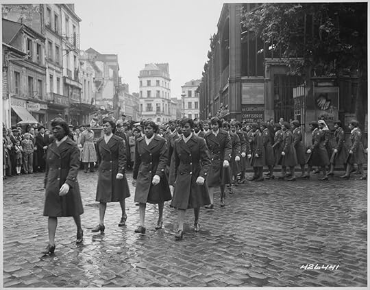Black and white historical photograph of a group of women in military-style coats and hats marching in formation on a cobblestone street. Behind them, another group of women in similar uniforms is also marching. Crowds of civilians, including men, women, and children, line both sides of the street, watching the parade. The scene takes place in a European city with multi-story buildings, shops, and a large tree visible in the background. The atmosphere suggests a formal or ceremonial event, possibly during or after World War II.