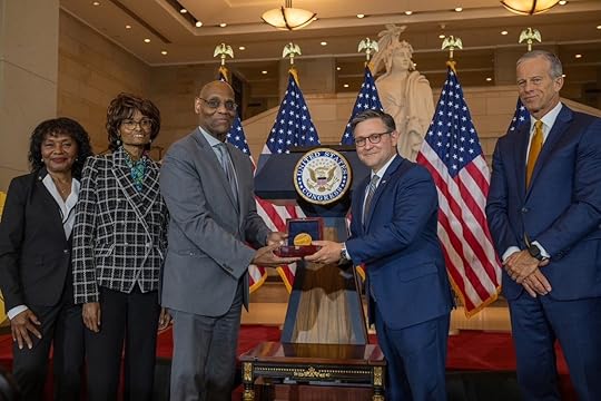 Five people in formal attire stand in a grand hall with four U.S. flags and a large statue in the background. Two men in the center hold a medal together in front of a podium displaying the U.S. House of Representatives seal.