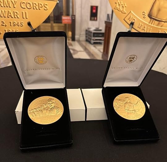 Two gold-colored commemorative medals in black velvet display boxes sit on a table. Each medal is engraved with images and text, with 'United States Mint' printed inside the box lids. In the blurred background are large medallions and a marble hallway.