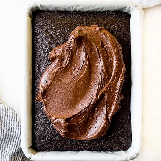 Thick, creamy chocolate buttercream frosting being spread onto a cool, freshly baked chocolate sheet cake in a parchment-lined pan, demonstrating a key step in a homemade cake recipe.