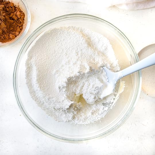 Confectioners' sugar (powdered sugar) being added to softened butter and cream cheese in a clear glass bowl with a spatula, a step in making homemade rich chocolate buttercream frosting.