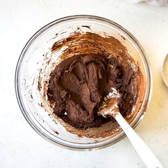 Partially mixed dark chocolate frosting in a clear glass bowl, showcasing the cocoa powder being incorporated into the creamy base with a white spatula, a key step in preparing a decadent homemade chocolate icing.