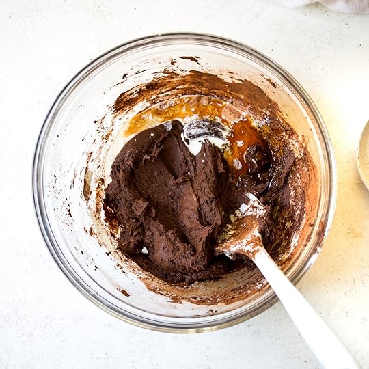 Overhead view of vanilla extract and heavy cream being poured into partially mixed chocolate frosting batter in a clear glass mixing bowl with a spatula, a liquid addition step for a smooth chocolate dessert topping.