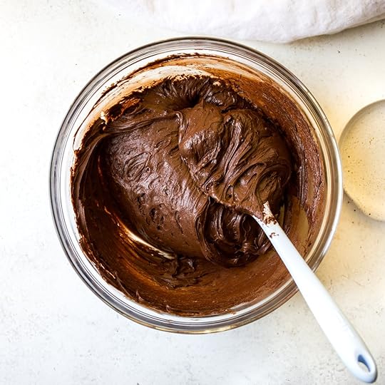 Finished and glossy chocolate rosting swirled in a clear glass bowl with a white spatula, perfectly blended for a decadent homemade dessert spread or cake topping.