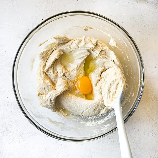 Raw egg yolk and white added to creamed butter and sugar mixture in a clear glass bowl with a spatula, a key step in preparing the cake batter for a homemade chocolate sheet cake recipe.