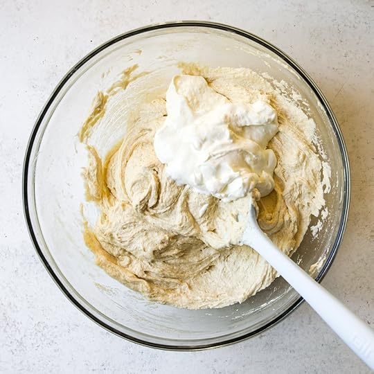 Sour cream being added to the creamed butter, sugar, and egg mixture in a clear glass bowl with a white spatula, a step in making moist cake batter for a homemade chocolate sheet cake recipe.