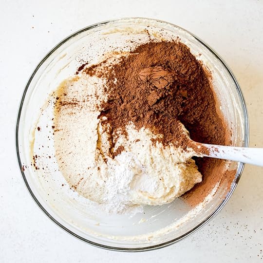 Unsweetened cocoa powder, baking powder, baking soda and salt added on top of wet cake batter ingredients in a clear glass bowl with a spatula, a key step in preparing the chocolate mixture for a homemade sheet cake.