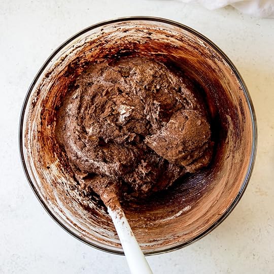 Partially mixed chocolate cake batter with visible streaks of cocoa powder and flour and lighter wet ingredients in a clear glass mixing bowl with a white spatula, illustrating a key step in preparing a homemade chocolate sheet cake.