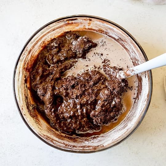 Liquid ingredients like milk and espresso being added to the thick chocolate cake batter in a clear glass mixing bowl with a white spatula, a crucial step for achieving a smooth consistency in a homemade chocolate sheet cake recipe.