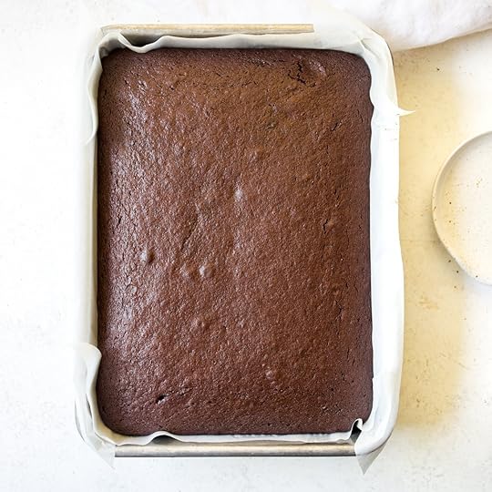 Freshly baked homemade chocolate sheet cake cooling in a parchment paper-lined rectangular baking pan, ready to be frosted, from an easy chocolate cake recipe.