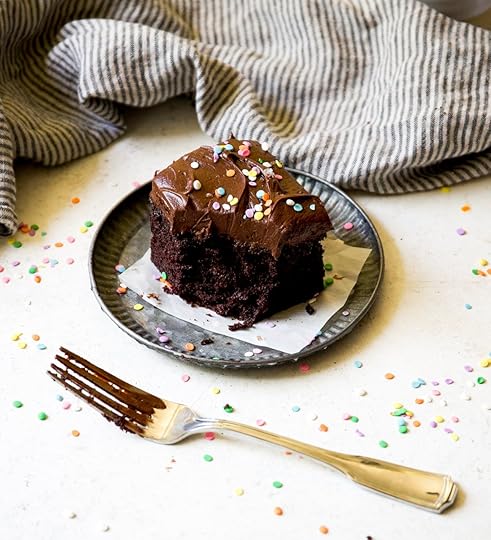 Single slice of moist chocolate sheet cake on a rustic metal plate, generously frosted with chocolate buttercream and topped with colorful rainbow sprinkles, with a chocolate-smeared fork nearby, ready to enjoy from a homemade cake recipe.
