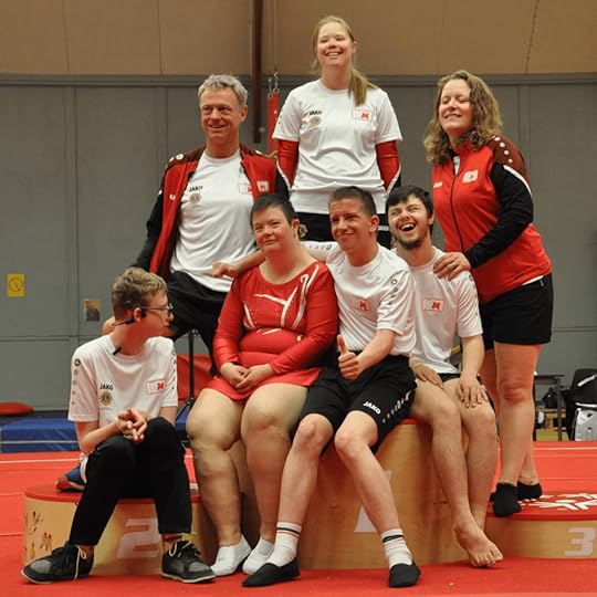 Groepsfoto van een gymnastiekteam, met atleten en coaches in sportoutfits, poseren op een podium. De atleten lijken te genieten van het moment en stralen blijdschap uit.