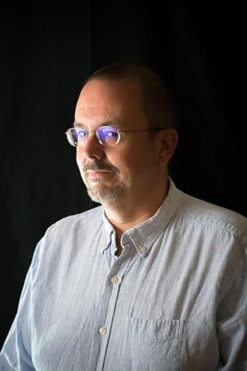 A man with short dark hair and a short silver beard faces you. He is wearing a white button-up shirt. Blue light reflects from his eye glasses.