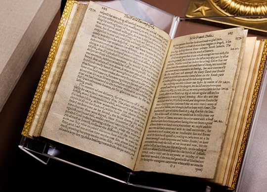 Medium photo of an open book resting in an acrylic holder. The pages are light brown with age, the ink of the printed copy still very black.