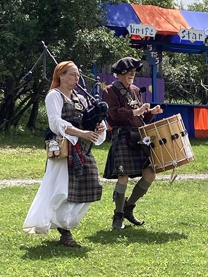 A woman in a tartan dress playing the bagpipes alongside a man in a brown outfit playing a drum, with a colorful festival backdrop.
