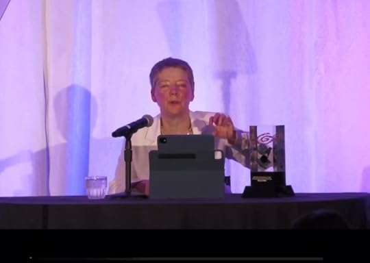 Screen shot of a video of a short-haired white woman in a white jacket on a well-lit stage. She is in a wheelchair at a table against a purple backdrop. Before her are an iPad on a stand, a microphone, and a large lucite column in which are embedded a variety of things. This is the award she is receiving.