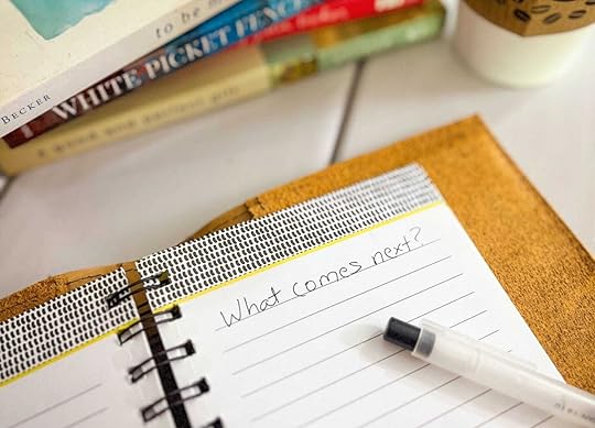 A close-up image of an open notebook with the handwritten words “What comes next?” on a lined page. A black-capped pen rests on the page. In the background, a stack of books is slightly out of focus, with one spine reading “WHITE PICKET FENCES” and another showing the author's name, “Becker.” A coffee cup with a sleeve decorated in a coffee bean pattern sits nearby.