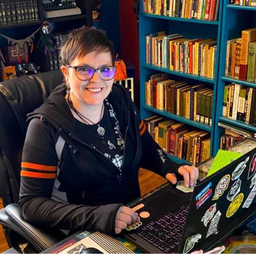 Portrait of author Jessica Crichton. A Caucasian woman smiles confidently. She has short brown hair and eye glasses, wears a dark hoodie. Before her a laptop is open and ready. Shelves of books line the walls behind her.