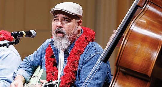 Musician wearing a red flower lei and a flat cap sings and plays guitar into a microphone during a performance. A double bass is visible beside him, suggesting a live music setting.