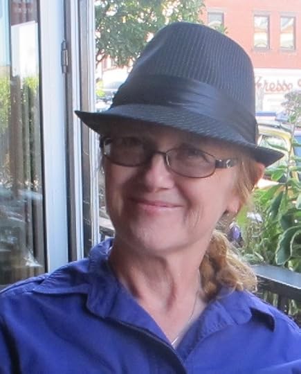 Portrait of author Joyce Reynolds-Ward. A caucasian woman smiles confidently. She is wearing a dark gray fedora and a navy blue open collared shirt.