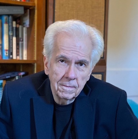 Portrait of author Gregory Frost. An older Caucasian man with white hair and a blue suit. His expression is somber.