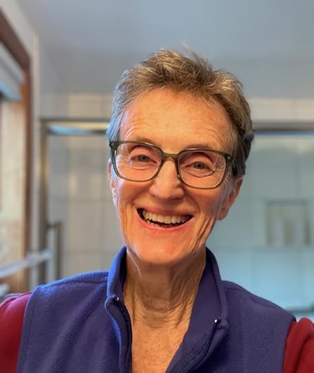 A smiling older woman with short gray hair and glasses is wearing a blue vest over a red shirt, standing in a brightly lit bathroom.