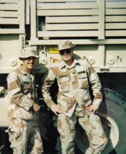 Two U.S. Army soldiers in desert camouflage uniforms standing in front of a large Army truck