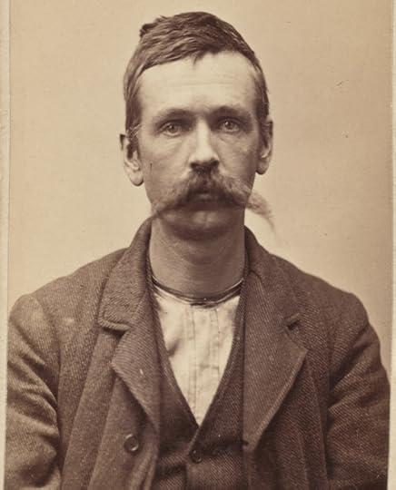 Sepia-toned, head and shoulder portrait shows a tired looking man, staring at the camera. He has short brown hair and a long handlebar moustance. His coat, vest and shirt appear well worn.