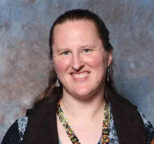 Author Sanan Kolva. A Caucasian woman smiles confidently. She has long brown hair and wears a rich cloak.