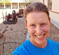 A white woman with short blond hair smiles. In the background is old mining equipment.