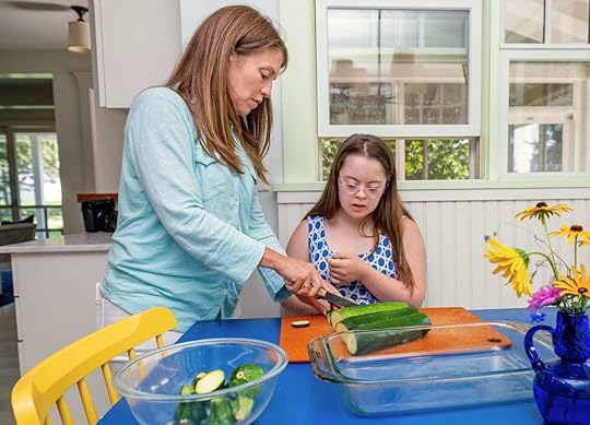 Penny and I are preparing food together in a kitchen. I'm showing Penny how to slice zucchini on a cutting board placed on a blue table. A glass baking dish and a bowl of chopped zucchini are nearby