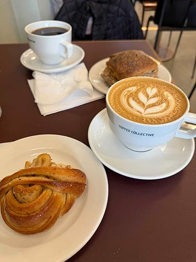 A cozy café table at Coffee Collective Bernikow in Copenhagen, featuring a cappuccino with elegant latte art, a black coffee, a cardamom bun, and a sandwich on rustic bread.