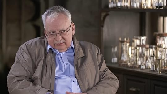 Andrzej Sapkowski in a brown coat and glasses sitting in a chair