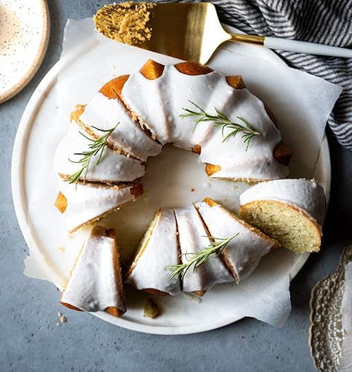 Overhead view of a whole vanilla bundt cake with white glaze and fresh rosemary, sliced on a white marble serving platter