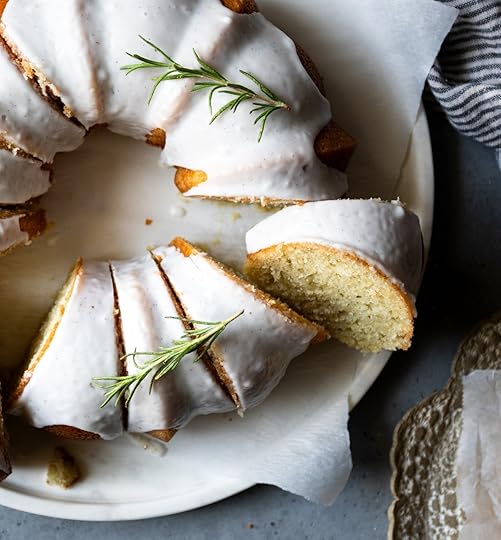 Overhead view of a whole vanilla bundt cake with white glaze and fresh rosemary, sliced on a white marble serving platter.