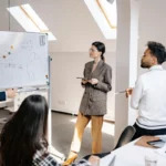 A well-dressed group engages in a brainstorming session, analyzing charts and data displayed on a whiteboard in a modern office space.