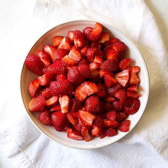 A white bowl filled to the brim with fresh, vibrant red strawberries, mostly halved or quartered, sitting on a white linen cloth.